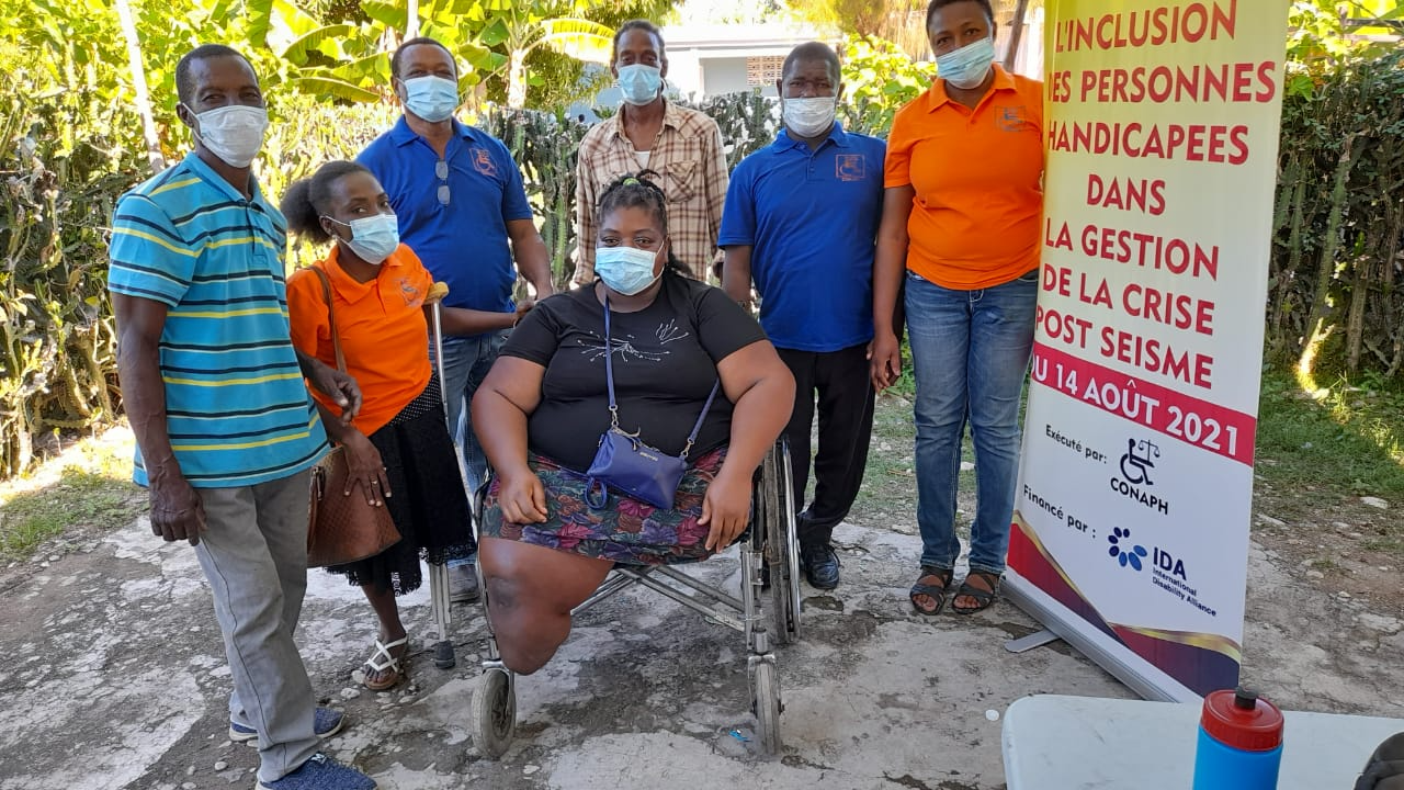 Photo of a group of people at a project site in Haiti. One of them in the centre is a wheelchair user along with some project workers and one person who is an amputee.